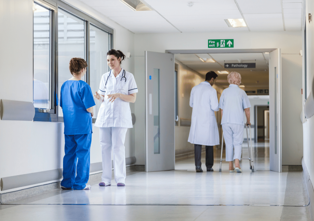 Medical professionals in scrubs and white coats converse and walk through a bright hospital hallway with white walls, large windows, and a green fire exit sign visible above a doorway marked "Pathology."