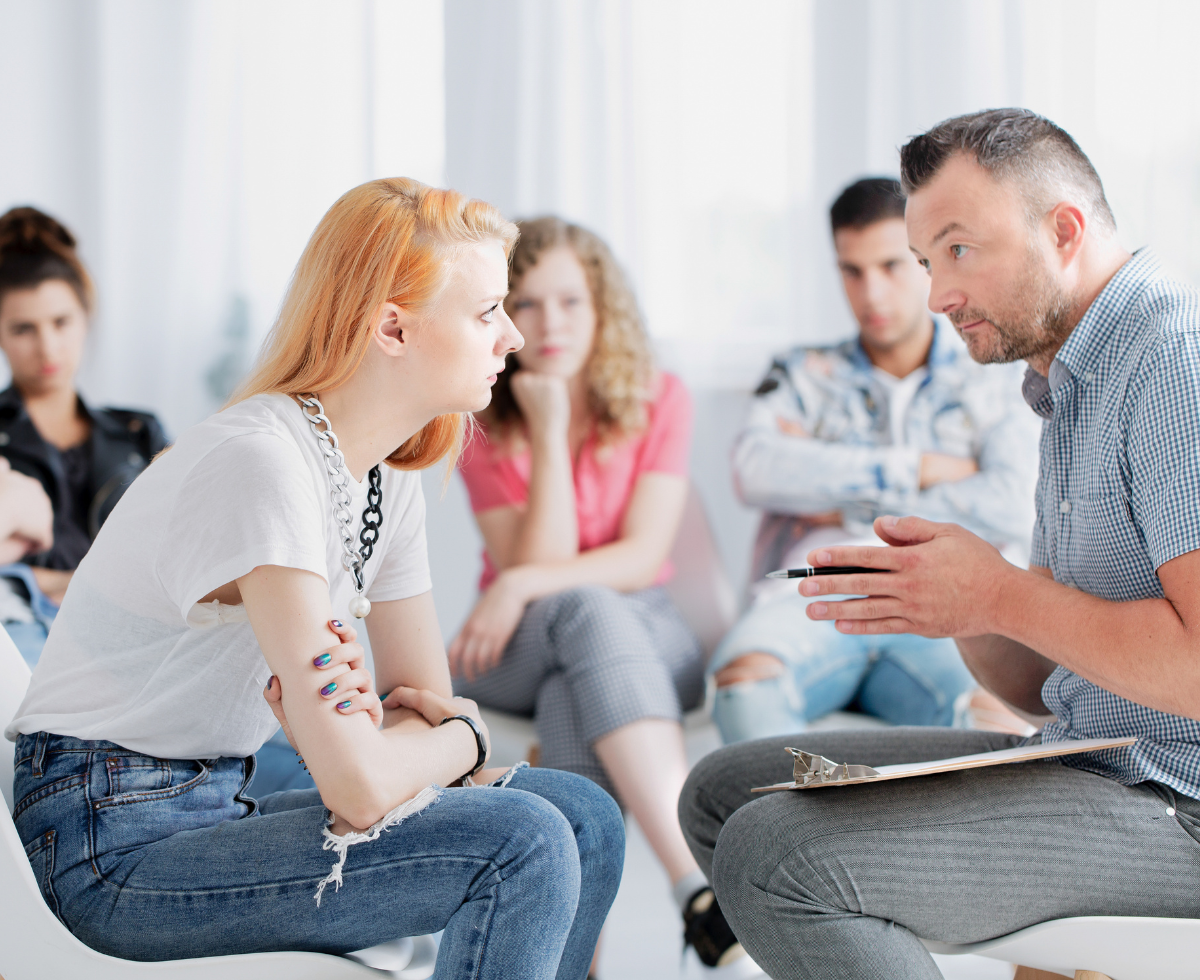 A therapist or counselor with a clipboard speaks with a young woman with blonde hair in a group therapy session, while other participants sit in the background forming a circle.