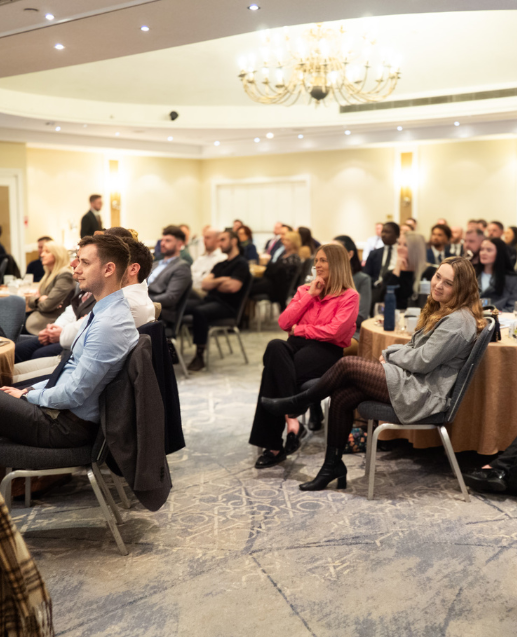 A business conference or seminar taking place in an elegant hotel ballroom, with professionally dressed attendees seated in rows listening to a presenter at the front of the room. The venue features ornate lighting, decorative ceiling details, and formal carpeting typical of upscale event spaces.