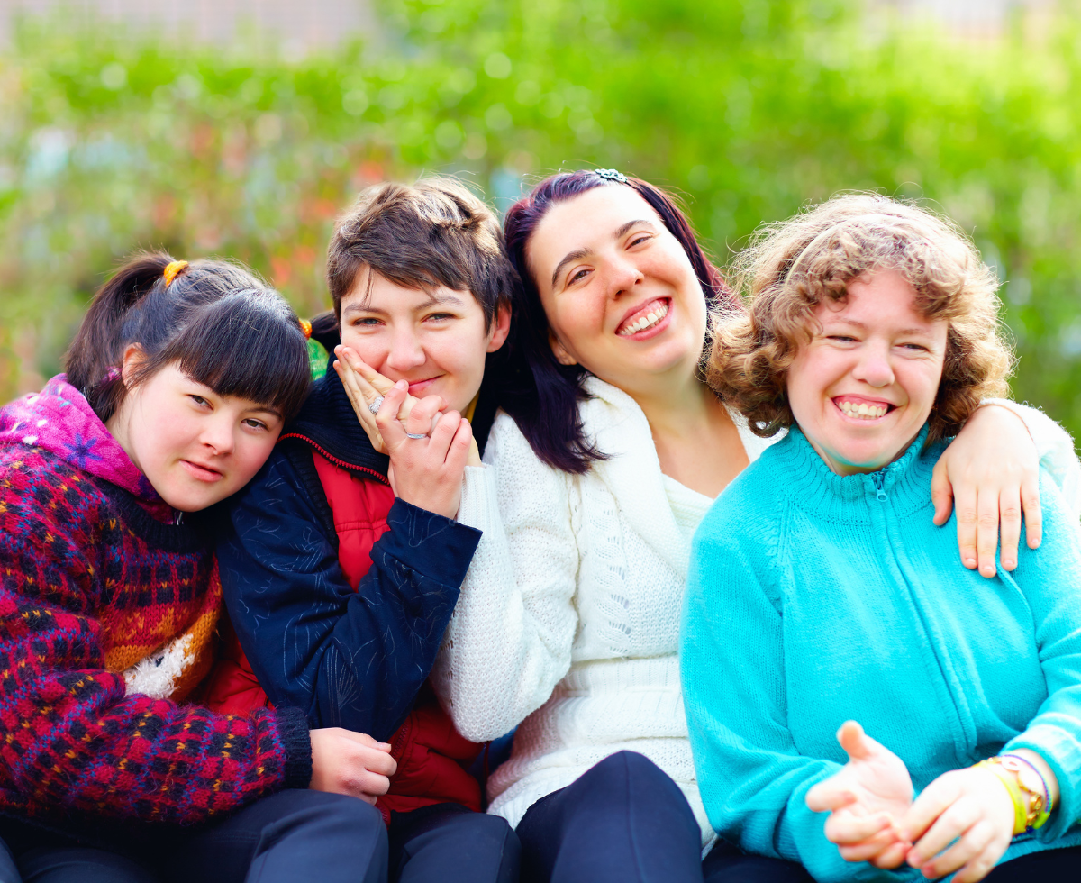 Five young people with developmental disabilities sit together outdoors, smiling and embracing each other in a warm group hug against a blurred green background of trees and foliage.