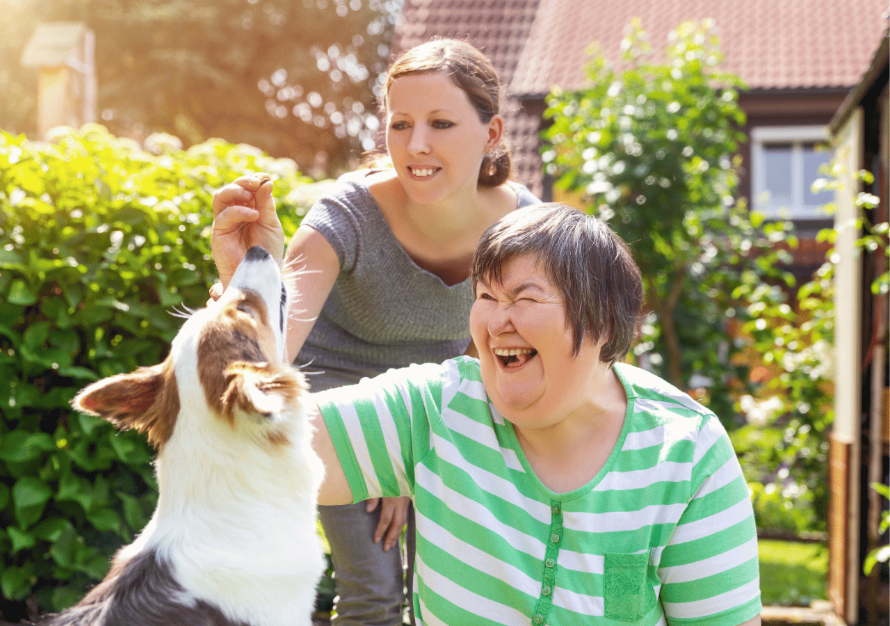 Two women enjoy a sunny outdoor moment with a small white and brown dog, with the woman in a green striped cardigan laughing joyfully as the other woman in gray holds the dog's paw. The scene takes place in a residential garden setting with lush greenery and traditional houses in the background.