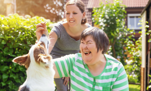 A caregiver assists a woman with developmental disabilities as they interact with a friendly dog outdoors in a sunny residential garden setting, both women smiling joyfully during the pet therapy session.