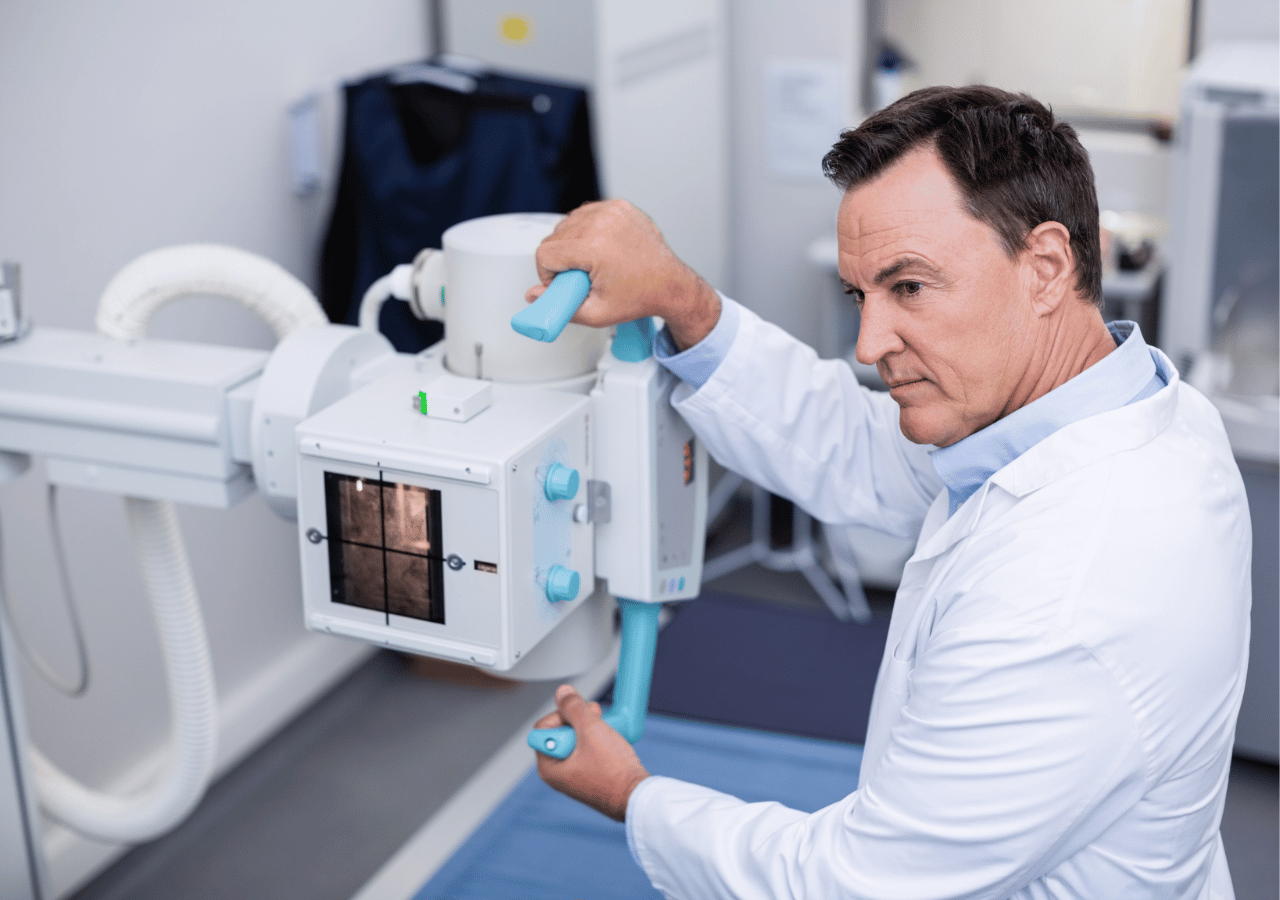 A medical professional in a white coat operates an X-ray machine in a clinical setting, adjusting the positioning controls while viewing an X-ray image displayed on the equipment's screen.