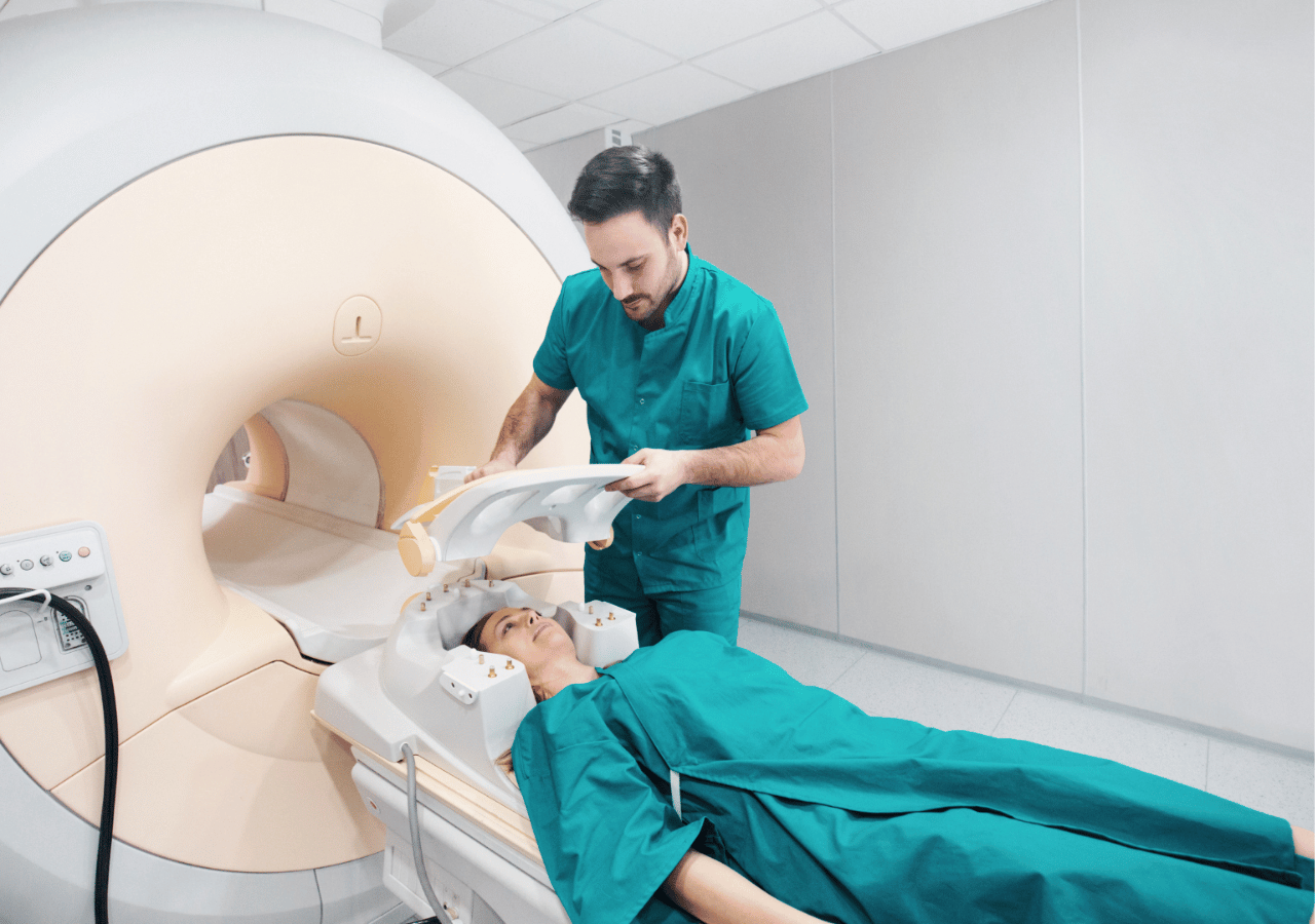A medical technician in green scrubs assists a patient lying on an MRI machine table, preparing for or monitoring a magnetic resonance imaging scan in a modern medical facility.