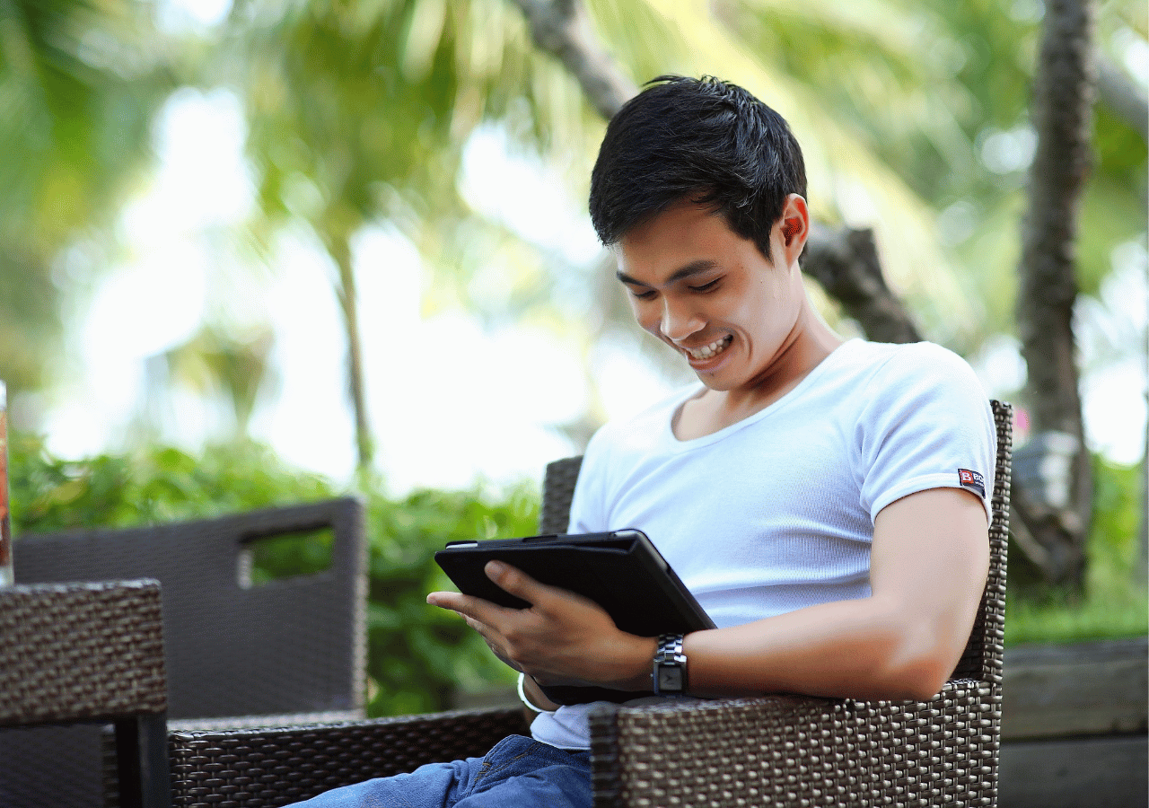 A young man in a white t-shirt sits in a wicker chair outdoors, smiling as he looks down at a tablet device in his hands, with lush green trees visible in the blurred background.