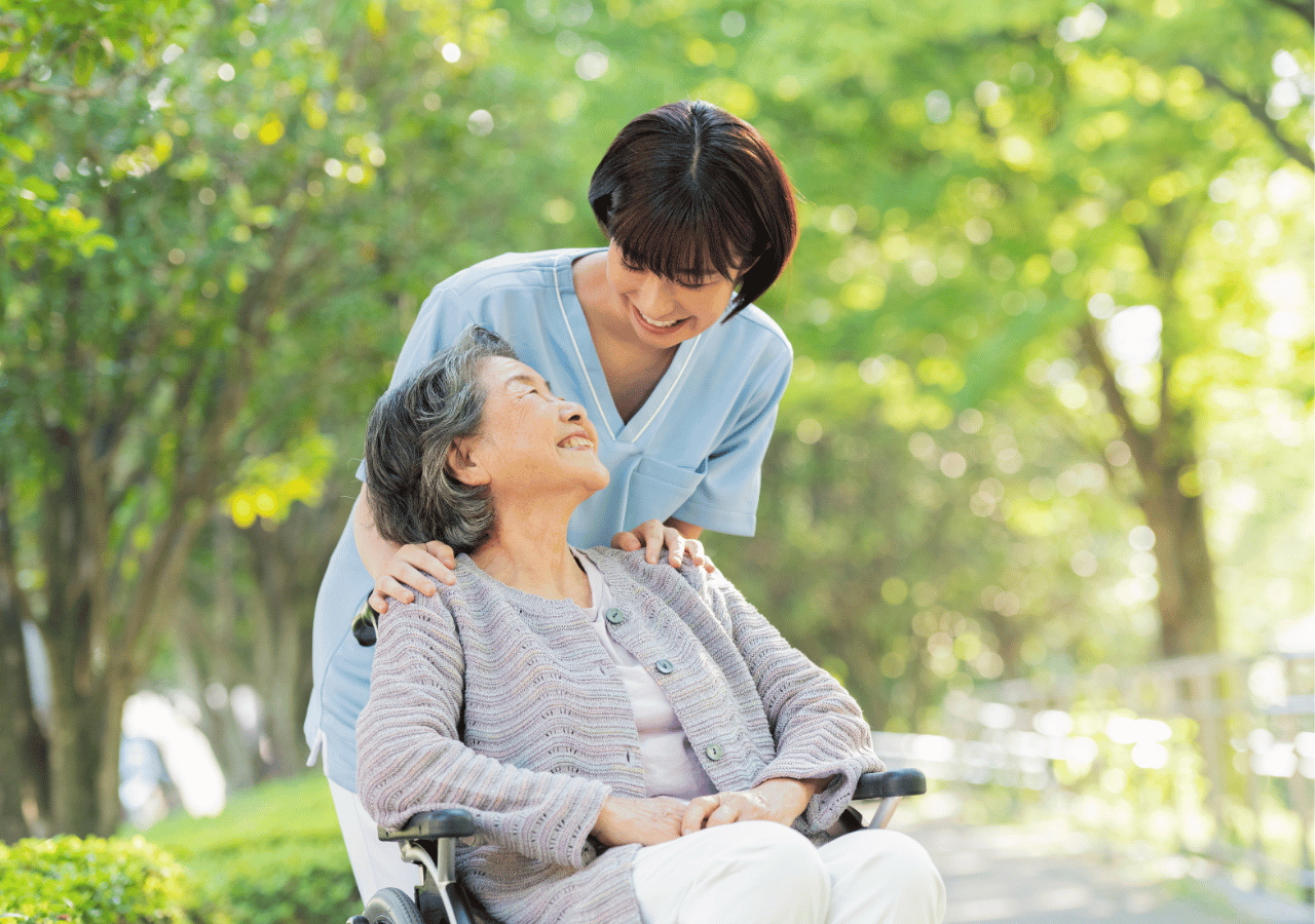A healthcare worker in light blue scrubs stands behind an elderly woman in a wheelchair, gently placing supportive hands on her shoulders as they share a warm moment together in a sunny, tree-lined outdoor setting.
