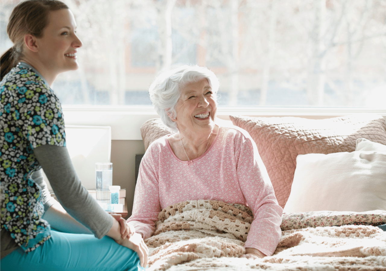 A healthcare worker in colorful scrubs sits beside an elderly woman in a pink sweater who is lying in bed, both smiling warmly as they hold hands in what appears to be a bright, comfortable care facility room.