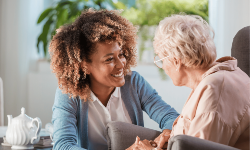 A young healthcare worker with curly hair smiles warmly while holding hands with an elderly woman with glasses in a bright, plant-filled room. The caring interaction suggests a compassionate moment between a caregiver and patient or resident.