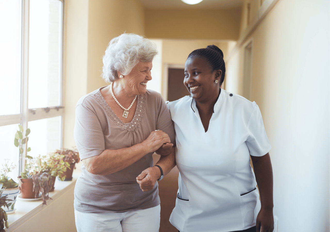 An elderly woman with white hair and pearl necklace walks arm-in-arm with a smiling healthcare worker in white scrubs down a bright hospital or care facility hallway.