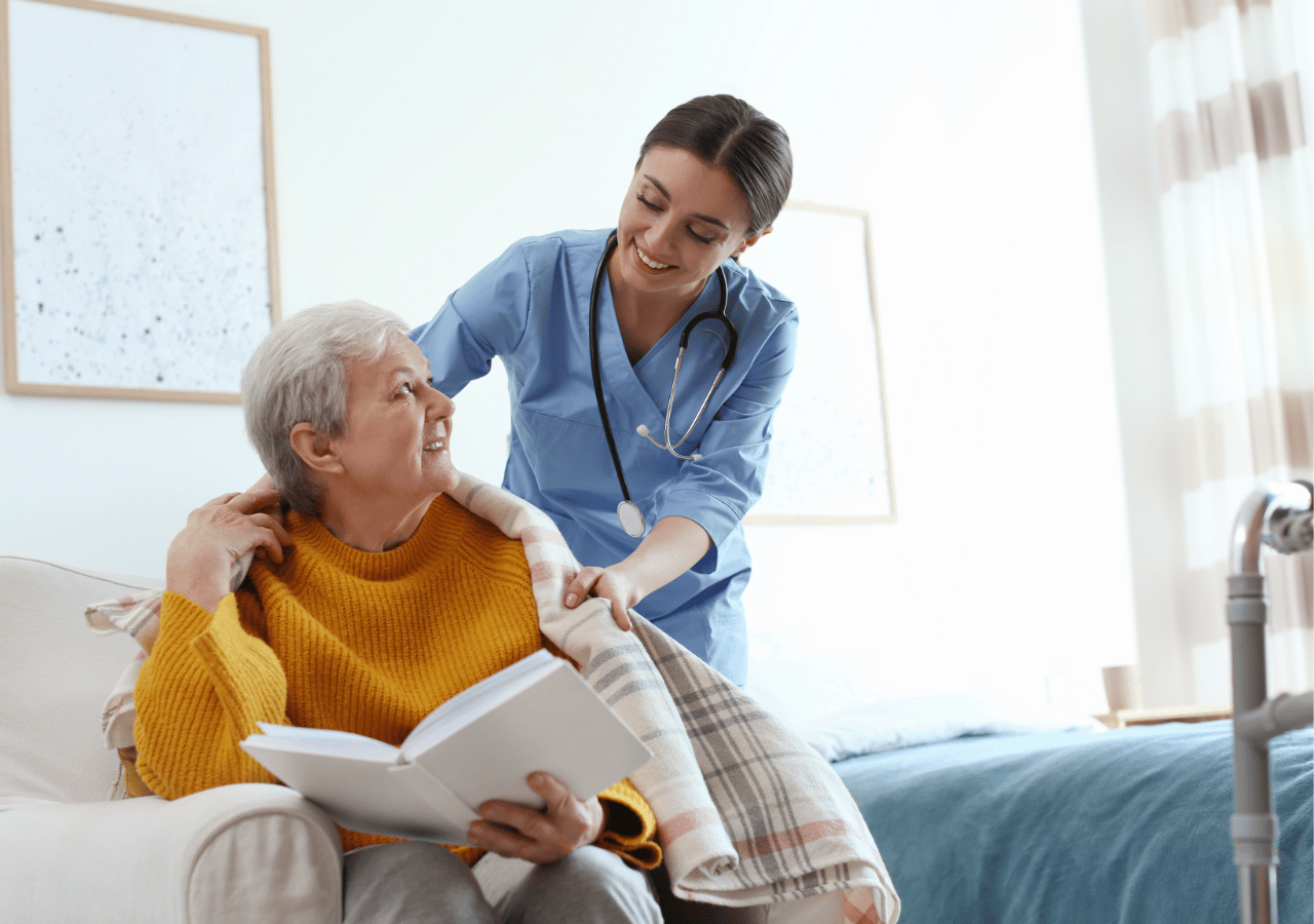 A healthcare worker in blue scrubs with a stethoscope provides gentle shoulder support to an elderly woman in a yellow sweater who is reading a book while sitting on a bed in a bright medical facility room.