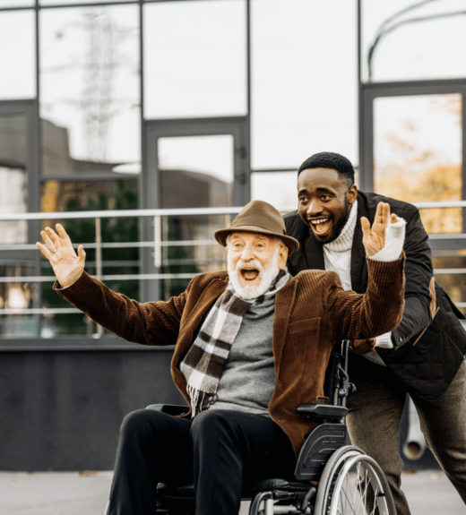 An elderly man in a wheelchair wearing a hat and scarf waves enthusiastically while a younger man stands behind him with his hands on the wheelchair handles, both smiling broadly on what appears to be an outdoor terrace or balcony with modern architecture in the background.