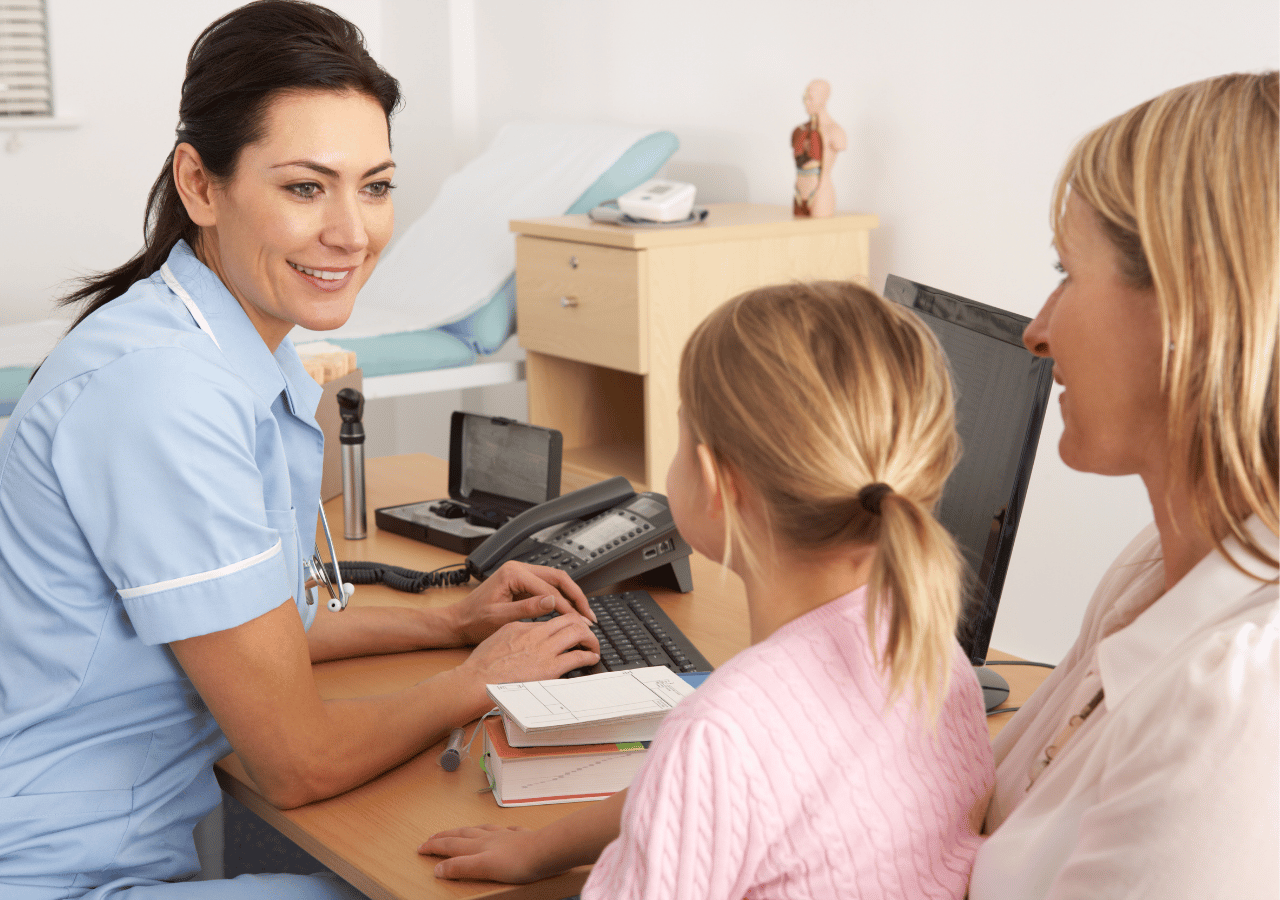 A healthcare professional in blue scrubs sits at a desk with a computer, consulting with a mother and young daughter in what appears to be a medical office or clinic setting.