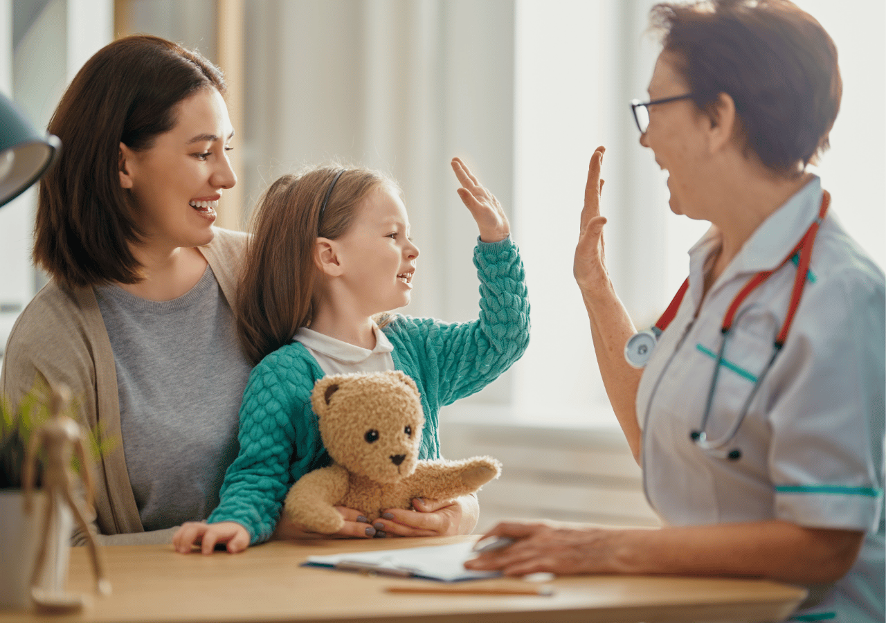 A young girl in a teal sweater gives a high-five to a female doctor while sitting next to her mother during a medical appointment, holding a small teddy bear.