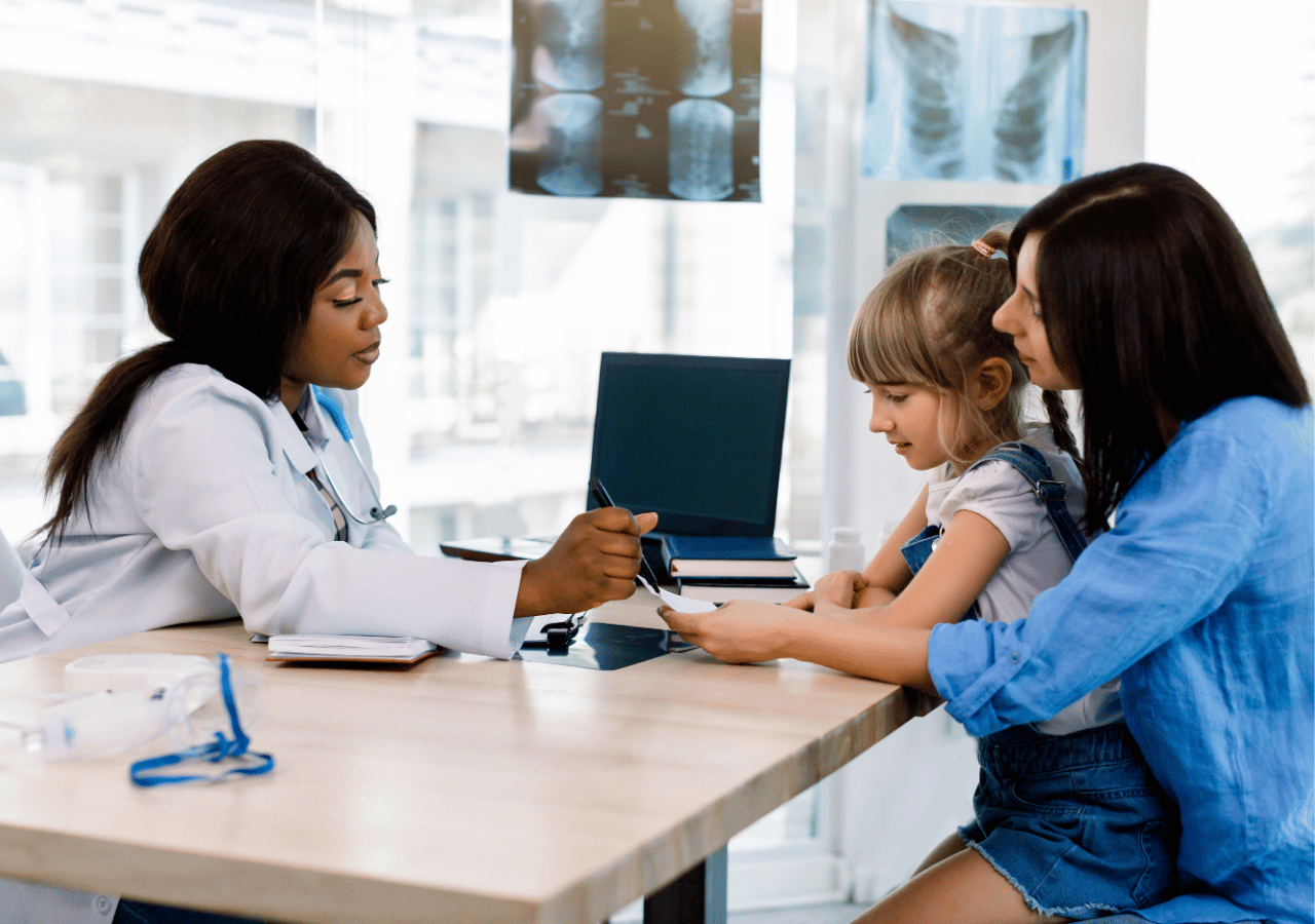 A female doctor in a white coat examines a young girl's arm while the child's mother sits beside her in a bright medical office with X-rays visible on the wall.