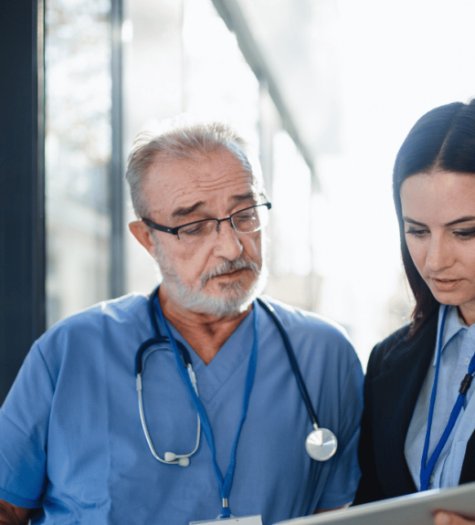 A senior male doctor in blue scrubs with a stethoscope discusses information on a tablet with a younger female colleague in business attire, both standing near a window in what appears to be a medical facility.