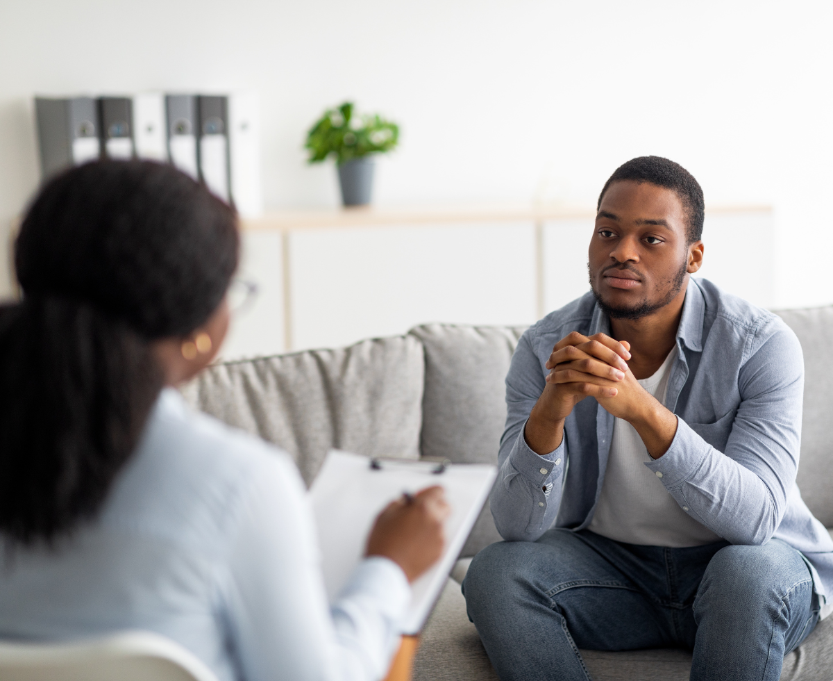 A young Black man in a light blue shirt sits on a gray couch with his hands clasped, speaking with a healthcare professional or counselor who is seated across from him holding a notepad in what appears to be a therapy or consultation session.