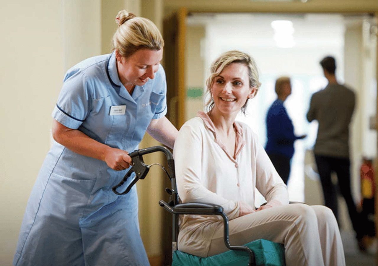 A healthcare worker in blue scrubs assists a smiling woman in a wheelchair in a hospital corridor, with other medical staff visible in the background.
