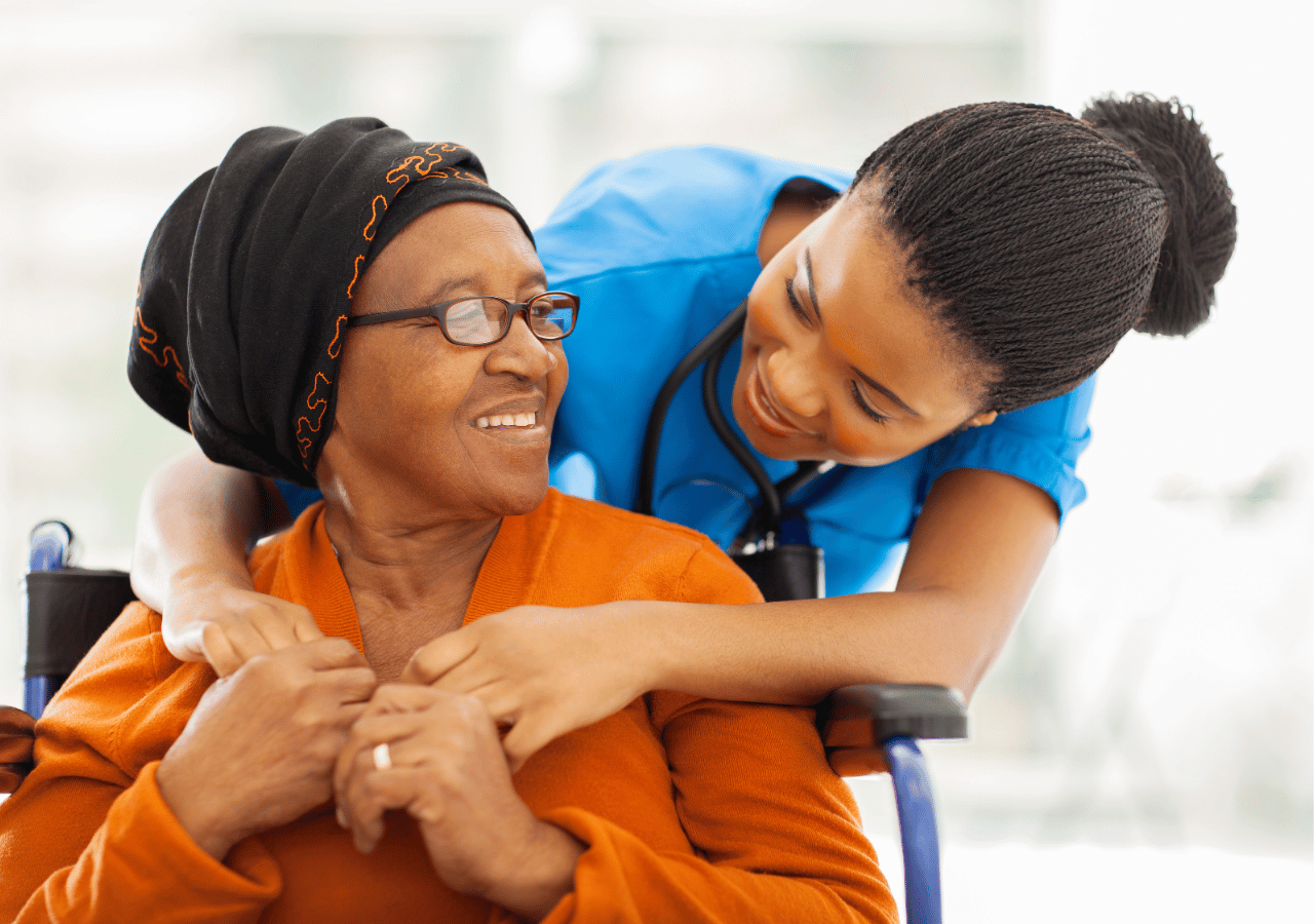 A healthcare worker in blue scrubs embraces an elderly woman wearing glasses and a head wrap who is seated in a wheelchair, both smiling warmly in what appears to be a medical or care facility.