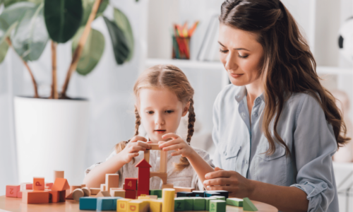 A woman in a light blue shirt sits with a young girl with braided hair as they play together with colorful wooden building blocks on a round table in a bright, plant-filled room.