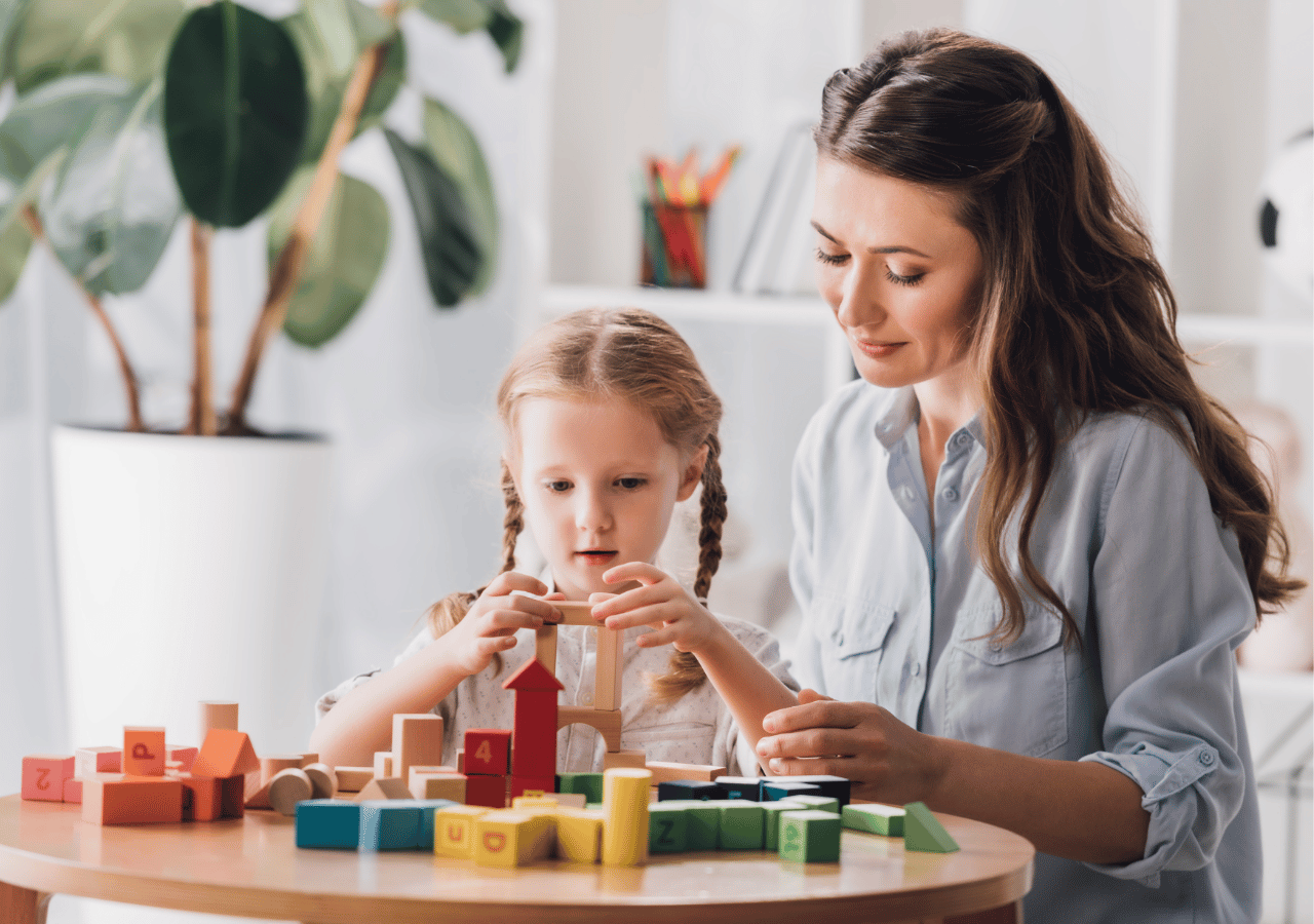 A woman in a light blue shirt sits with a young girl with braided hair as they play together with colorful wooden building blocks on a round table in a bright, plant-filled room.