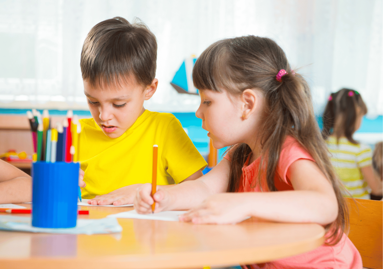 Two young children sit at a classroom table working on an art project together, with the boy in a yellow shirt and girl in a coral shirt using colored pencils and supplies from a blue container.