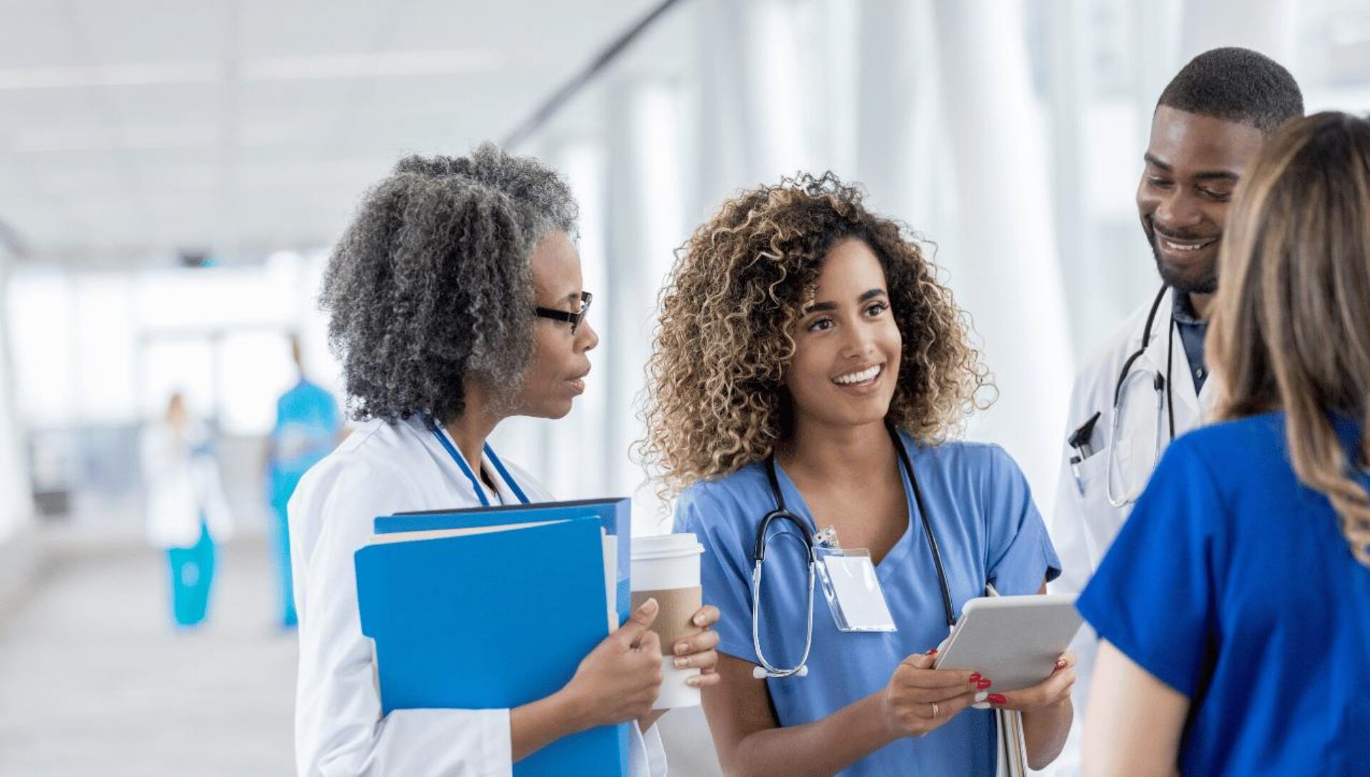 Four healthcare professionals in medical attire gather in a bright hospital corridor, with one holding a tablet and blue folder while they engage in discussion. The group includes doctors and nurses wearing scrubs and white coats with stethoscopes, appearing to collaborate on patient care.
