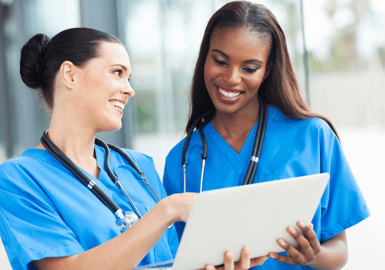Two healthcare professionals in blue scrubs with stethoscopes smile while reviewing documents together in a bright medical facility.