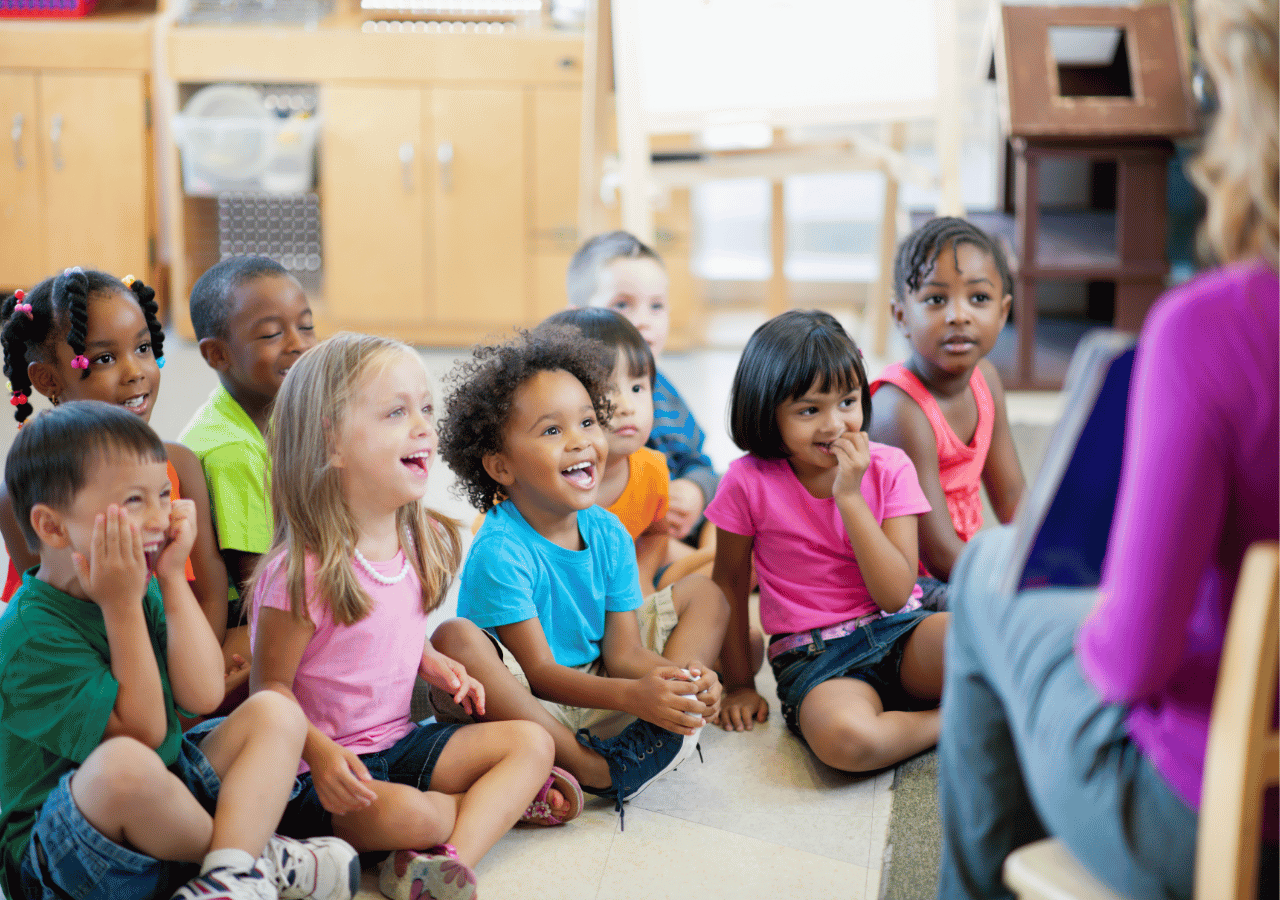 A diverse group of young children sits cross-legged on a classroom floor, smiling and looking attentively at someone in purple clothing who appears to be reading or teaching. The classroom setting includes wooden furniture and educational materials in the background.
