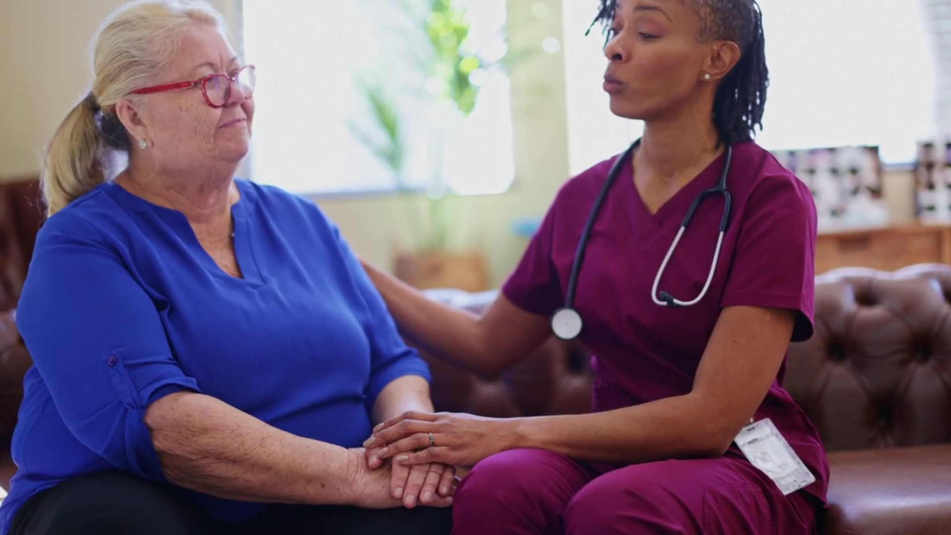 A healthcare worker in burgundy scrubs and stethoscope sits beside an elderly woman in blue clothing, holding her hands in a caring gesture during what appears to be a medical consultation or care visit.