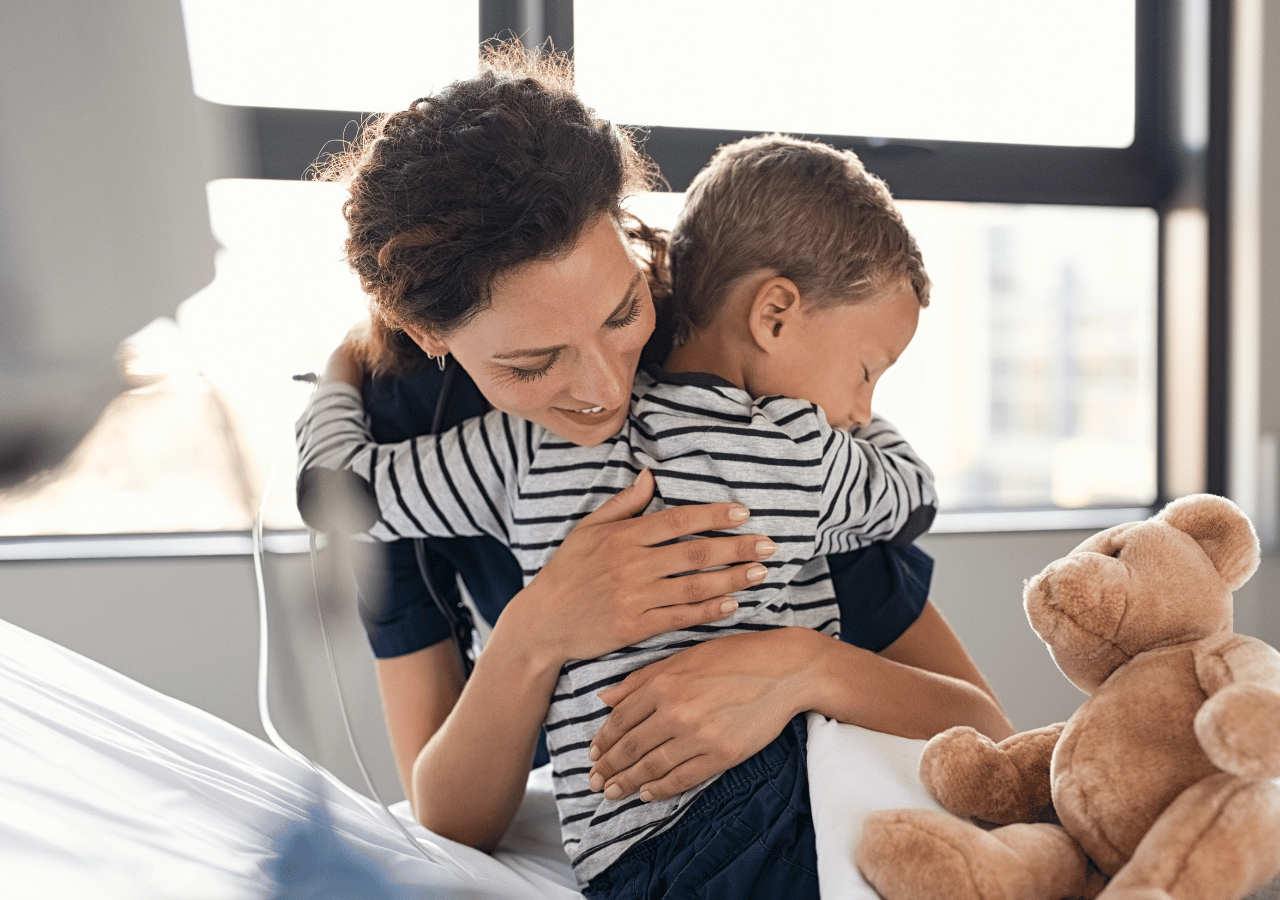 A mother with curly dark hair embraces her young son wearing a striped shirt as they sit together on a bed next to a teddy bear, with natural light streaming through windows behind them.