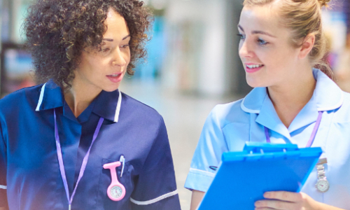 Two healthcare professionals in scrubs walk together through a hospital corridor, with one holding a blue clipboard while they appear to be discussing patient care or medical matters.