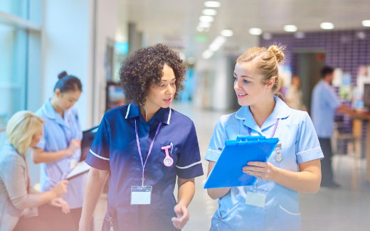 Two healthcare professionals in scrubs walk together down a hospital corridor, one holding a clipboard while they appear to be in conversation. Other medical staff can be seen working in the background of the busy healthcare facility.