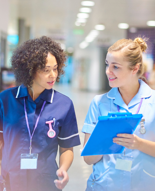 Two healthcare professionals in scrubs walk together through a hospital corridor, with one holding a blue clipboard while they appear to be discussing patient care or medical matters.