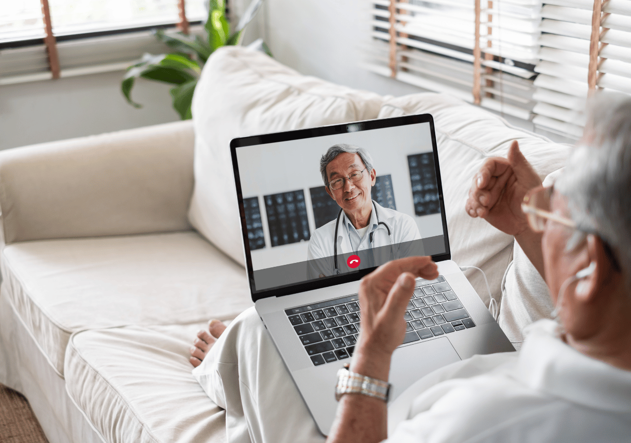 A person sits on a white couch having a video call consultation with a doctor wearing a stethoscope, who appears smiling on the laptop screen in what looks like a medical office setting.