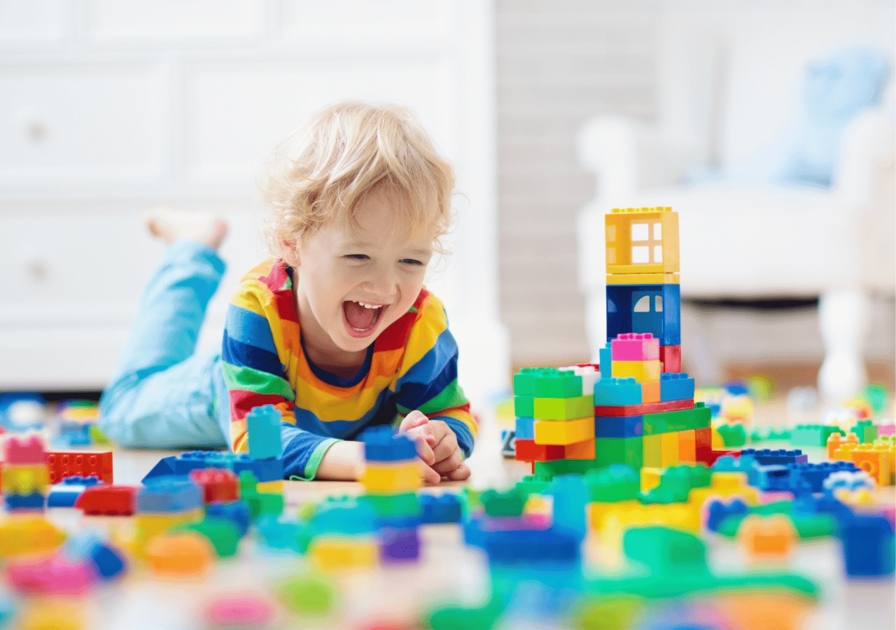 A young blonde child in a colorful striped shirt lies on their stomach, smiling joyfully while playing with an abundance of brightly colored building blocks scattered across the floor in what appears to be a playroom or home setting.
