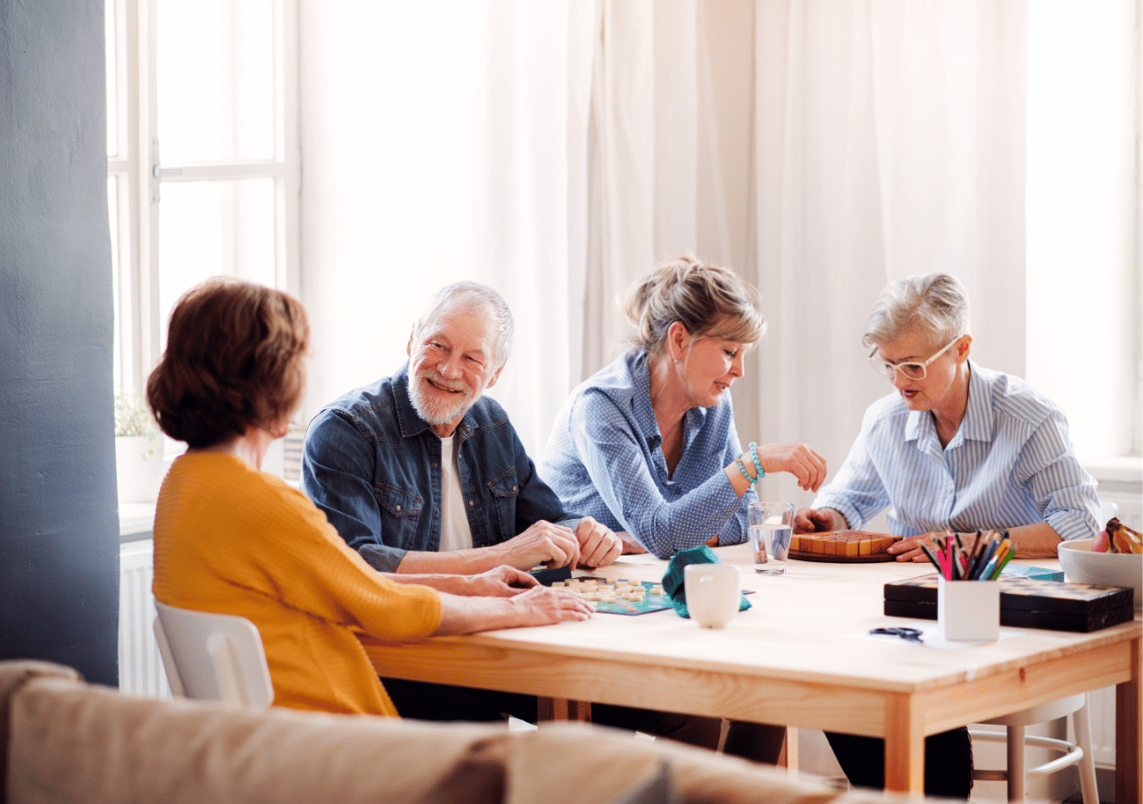 Four older adults sit around a wooden table engaged in a creative activity or game, with art supplies and materials spread across the surface in a bright, naturally lit room with large windows.