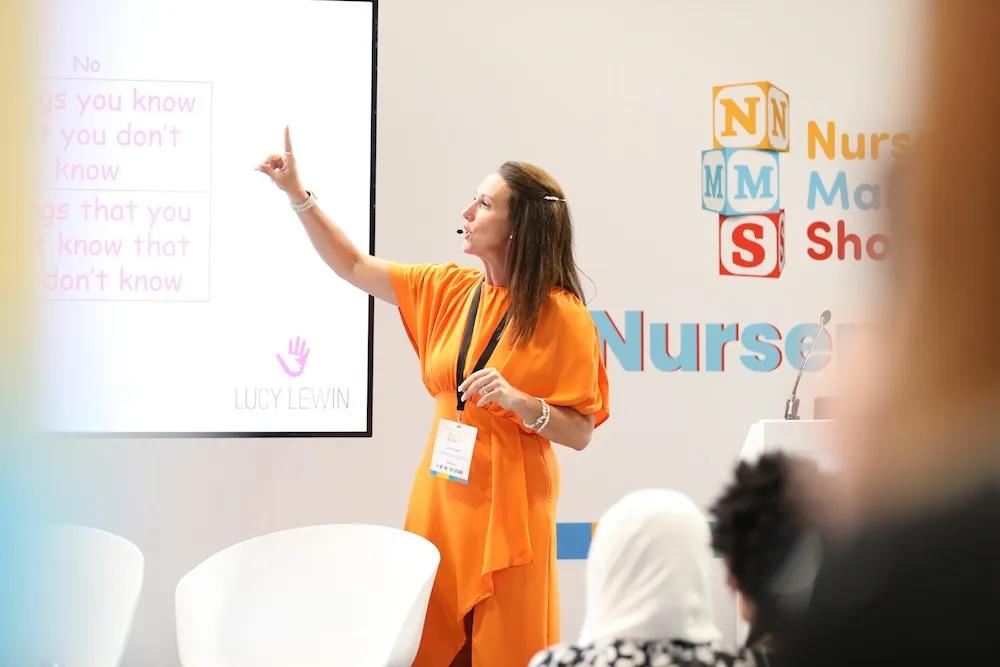 A woman in an orange outfit presents to an audience at what appears to be a nursing conference, pointing to a projected slide while standing near white chairs and promotional materials with nursing-related branding visible in the background.