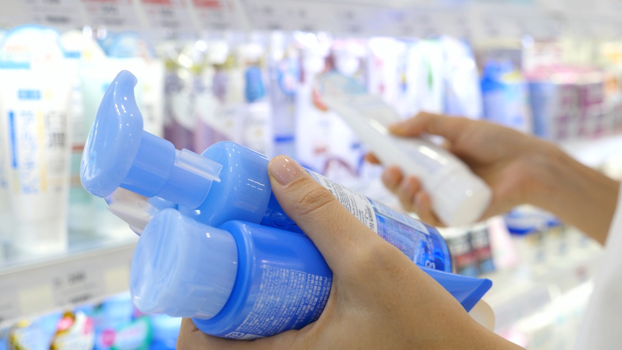 A person's hands hold blue nasal spray bottles while shopping in a pharmacy, with shelves of medical products visible in the blurred background.