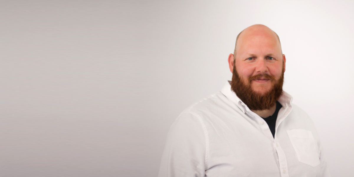 A bald man with a full brown beard wearing a white button-up shirt stands against a light gray background, smiling at the camera in what appears to be a professional headshot or portrait.