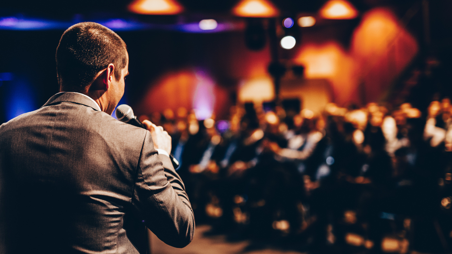 A speaker in a brown suit jacket holds a microphone while addressing a blurred audience in a dimly lit venue with warm overhead lighting.