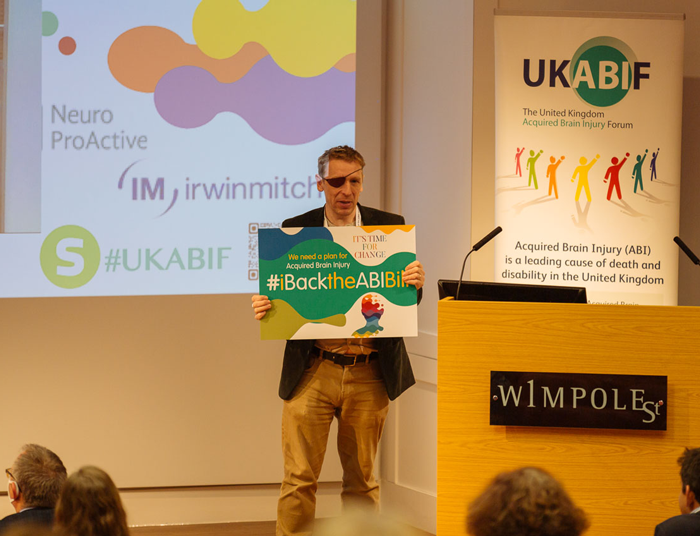A man stands at a podium holding a green "#BackTheABITeam" sign at a UKABIF (UK Acquired Brain Injury Forum) conference, with presentation screens showing Neuro ProActive and brain injury awareness information visible behind him.