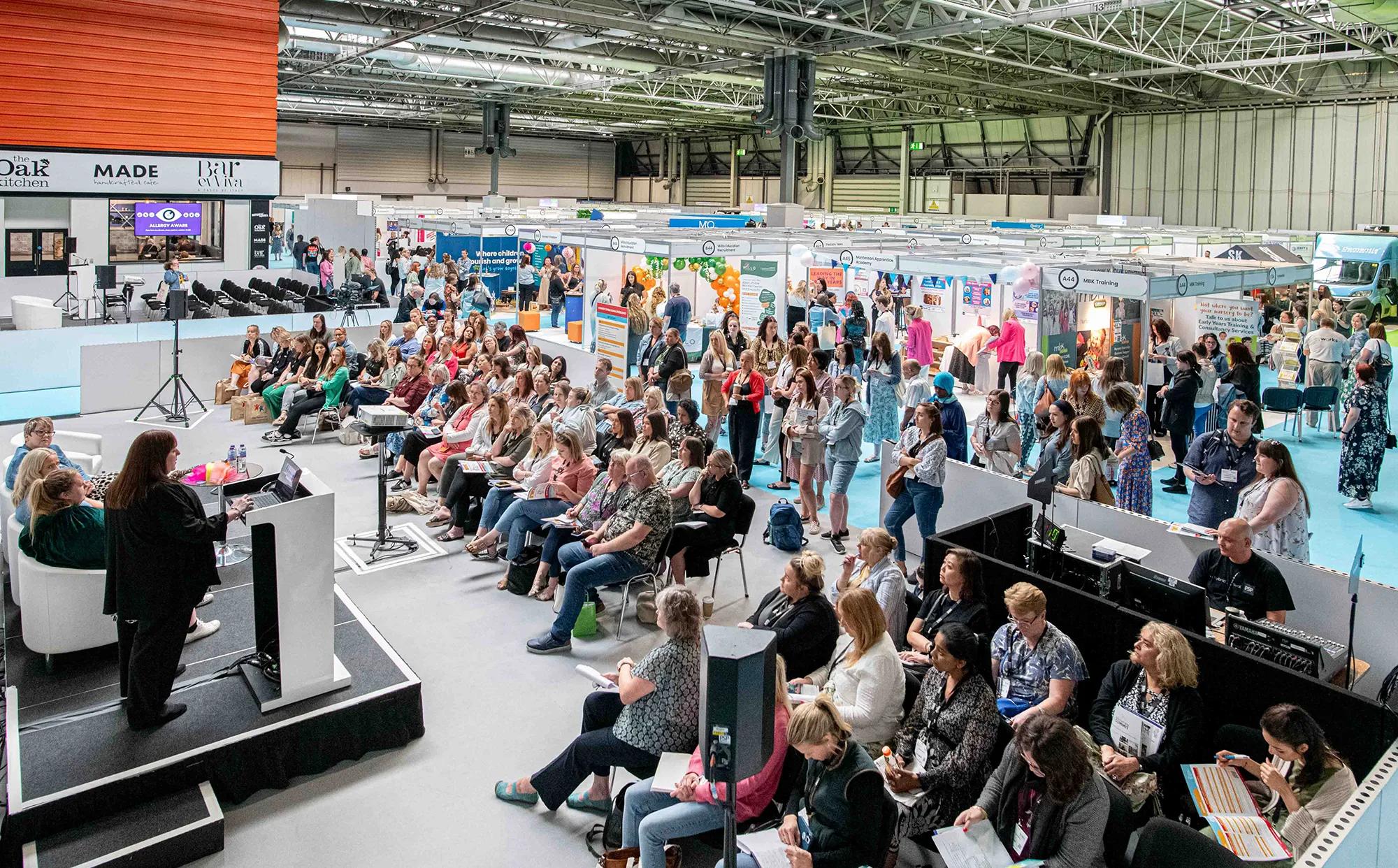A large indoor trade show or exhibition hall with hundreds of attendees seated in rows facing a presentation stage, while vendor booths and displays fill the background space under industrial ceiling lighting.