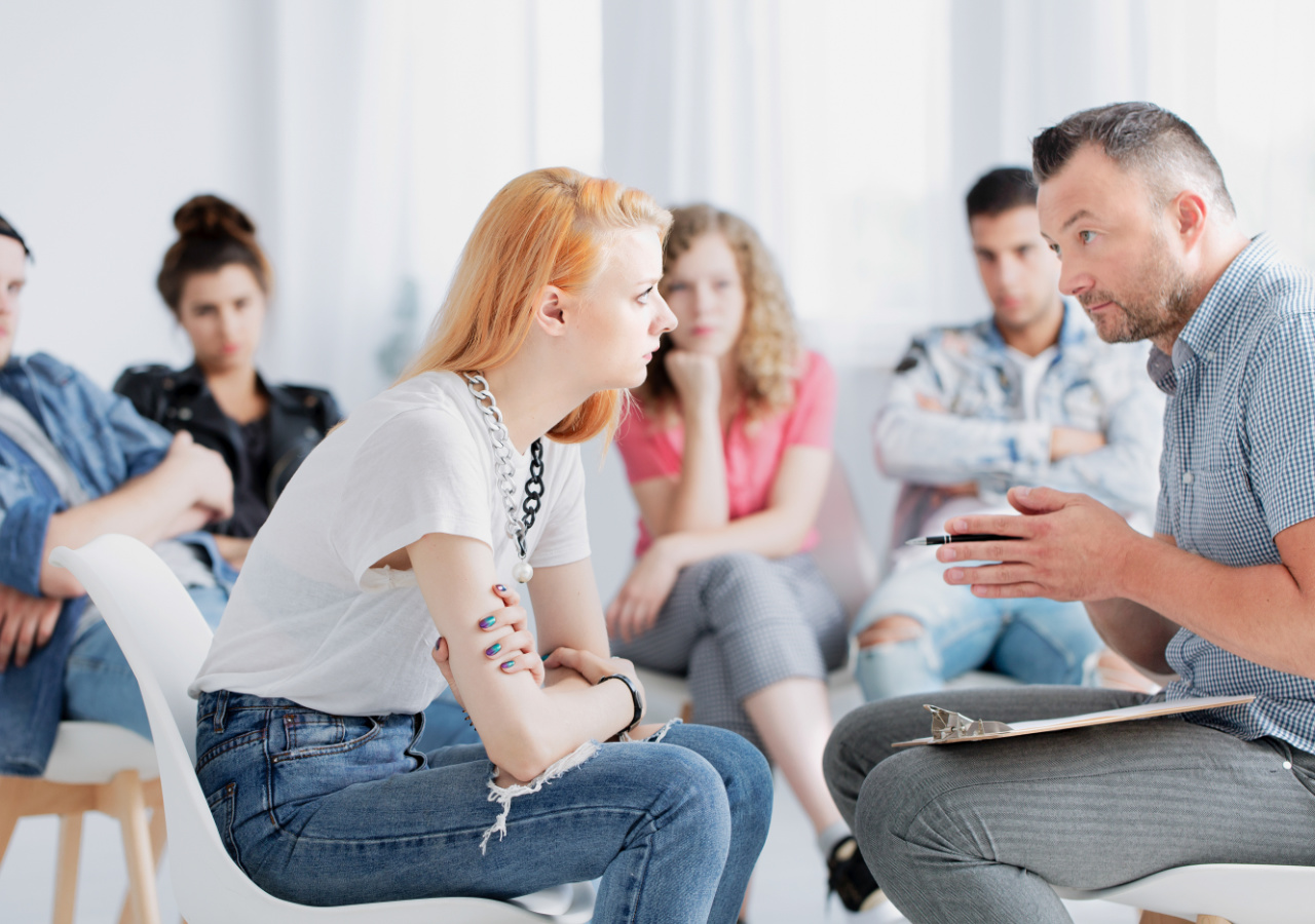 A therapist or counselor with a notebook sits facing a young woman with blonde hair and bandages on her arm during what appears to be a group therapy session, with other participants visible sitting in chairs in the background.