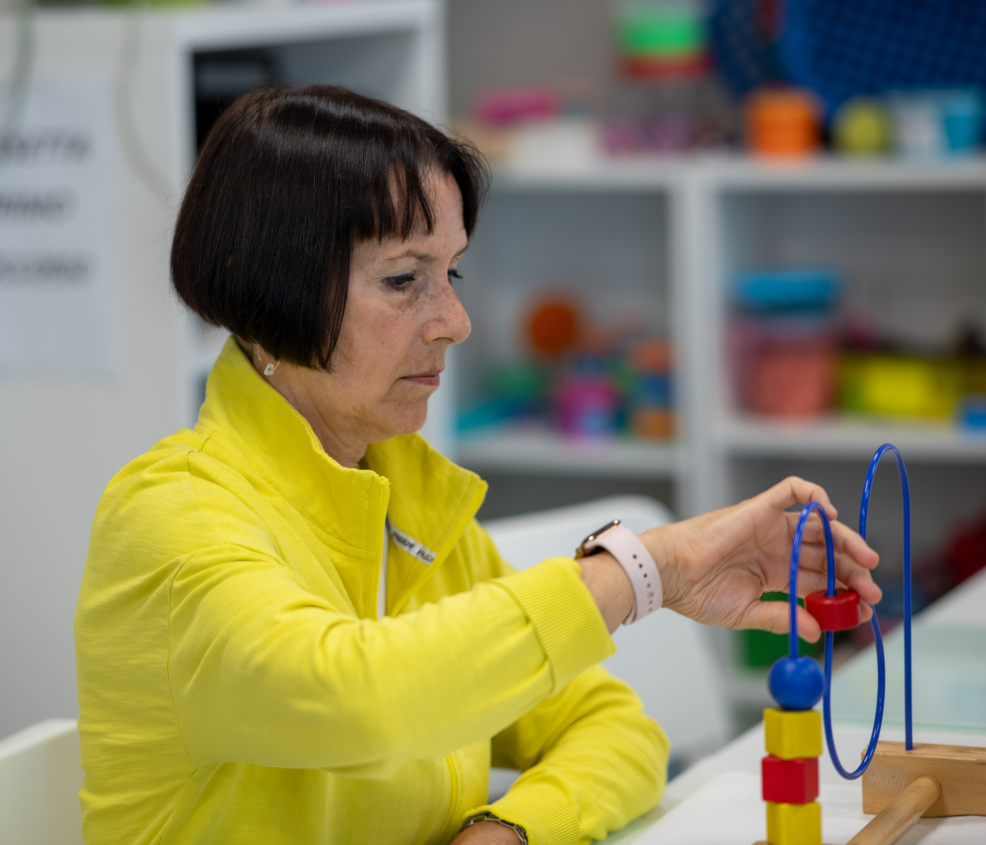 A woman in a bright yellow jacket concentrates while manipulating a colorful educational toy with beads and wire loops, seated at a table in what appears to be a therapy or learning environment with shelves of materials in the background.