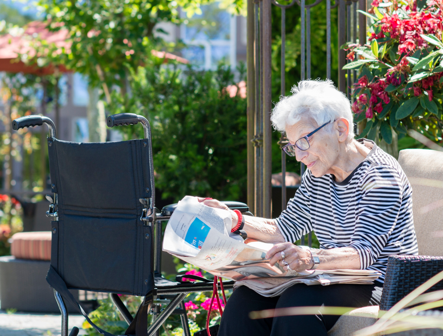 An elderly person with white hair and glasses sits outdoors reading a newspaper, with a wheelchair positioned nearby and lush garden greenery and flowers visible in the background.
