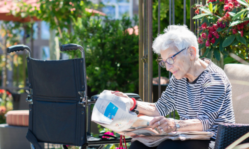 An elderly person with white hair and glasses sits outdoors reading a newspaper, with a wheelchair positioned nearby and lush garden greenery and flowers visible in the background.