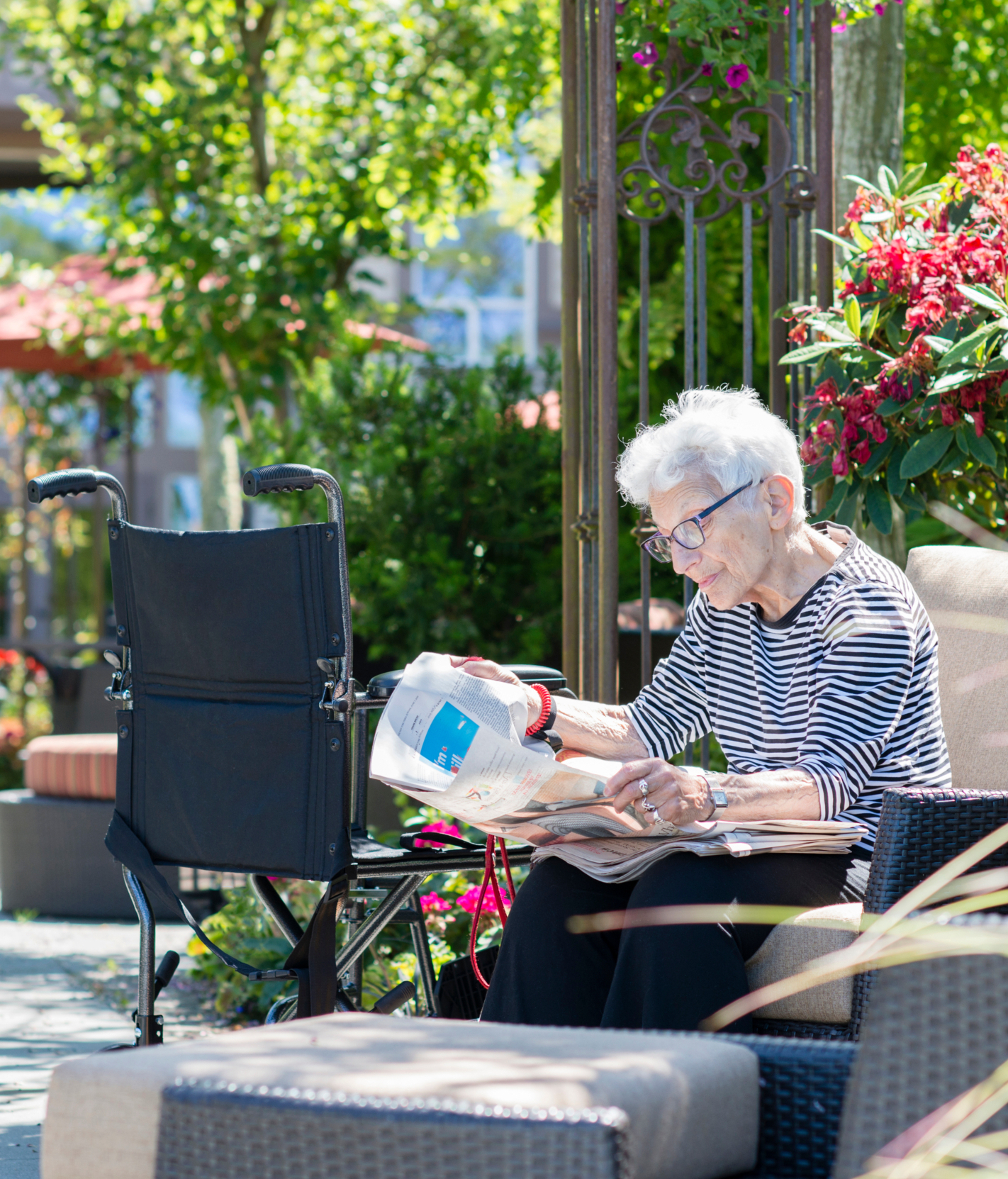 An elderly person with white hair and glasses sits outdoors reading a newspaper, with a wheelchair positioned nearby and lush garden greenery and flowers visible in the background.