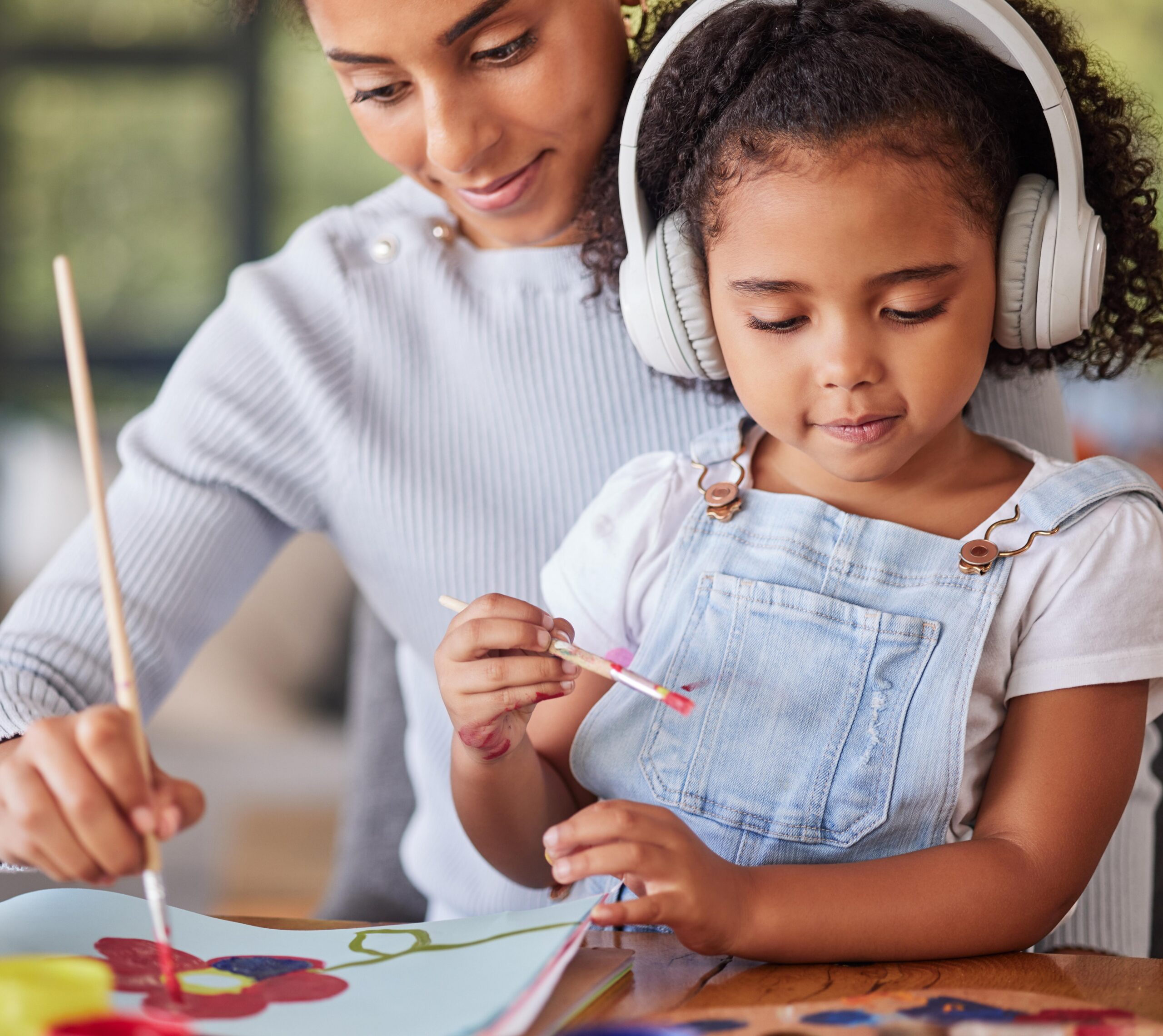 A young child wearing white headphones and a light blue denim overall sits at a table painting with watercolors while an adult woman in a white sweater sits beside them, both focused on the creative activity.