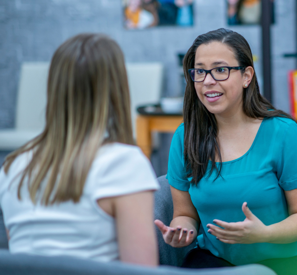 A woman wearing glasses and a teal shirt gestures with her hands while speaking to another person with blonde hair in what appears to be a professional or educational setting with shelving visible in the background.