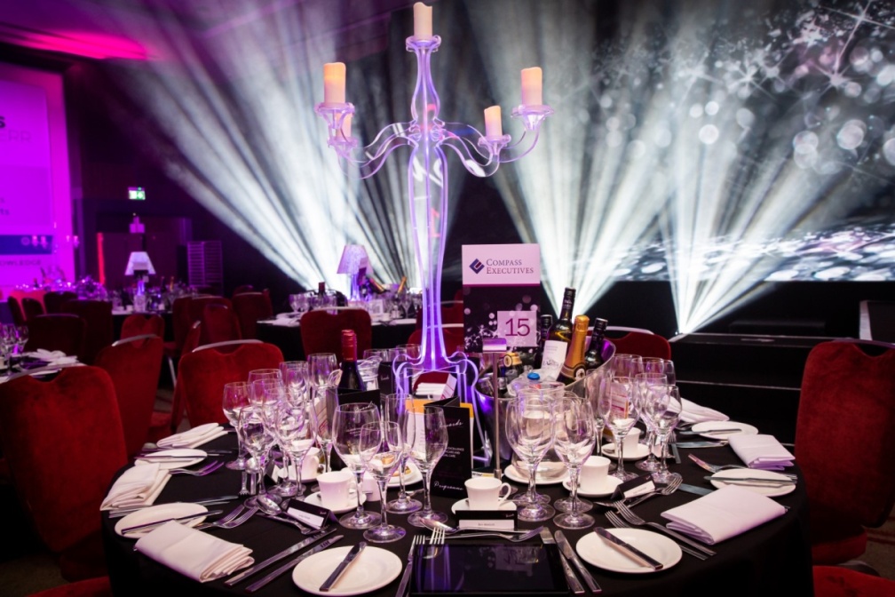 An elegant event venue with dramatic stage lighting featuring white beams and purple ambiance, showcasing a formal dining table with red chairs, crystal candelabra centerpiece, wine glasses, and place settings for what appears to be a corporate gala or awards dinner.